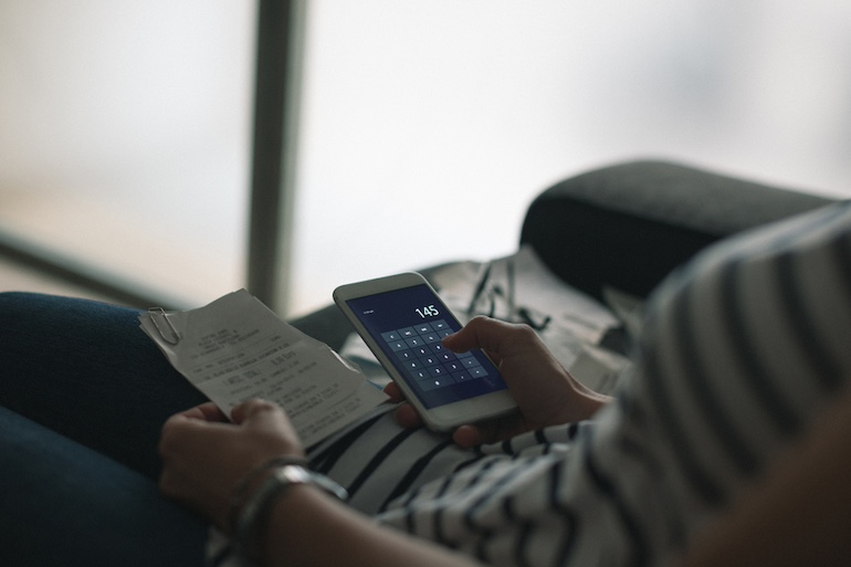 Person hat Dokumente in der einen und einen Smartphone Taschenrechner in der anderen Hand