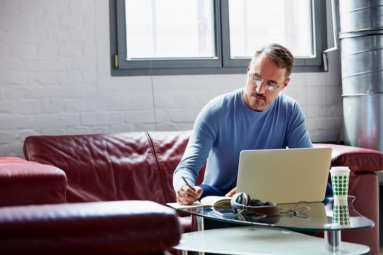 Vermieter mit Lesebrille sitzt auf dem Sofa am Laptop, er notiert in einen Block Zahlen.